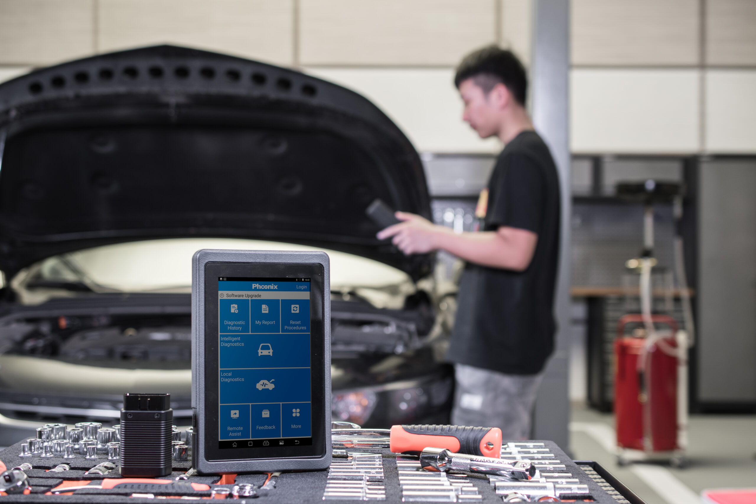 Giao diện chính máy chẩn đoán TOPDON Phoenix với các mục Diagnostic History, My Report, Reset Procedures, Intelligent Diagnostics, Local Diagnostics, Remote Assist, đặt trên bàn đồ nghề, kỹ thuật viên đứng cạnh xe mở nắp capo phía sau