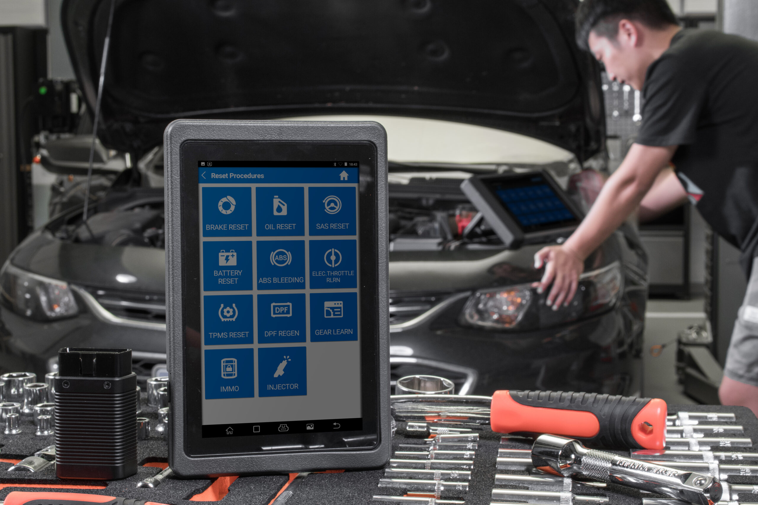 Màn hình Reset Procedures trên máy chẩn đoán TOPDON gồm Brake Reset, Oil Reset, SAS Reset, Battery Reset, ABS Bleeding, Elec Throttle Relrn, TPMS Reset, DPF Regen, Gear Learn, IMMO, Injector, đặt trên bàn đồ nghề trong xưởng, kỹ thuật viên đang thao tác trên xe phía sau